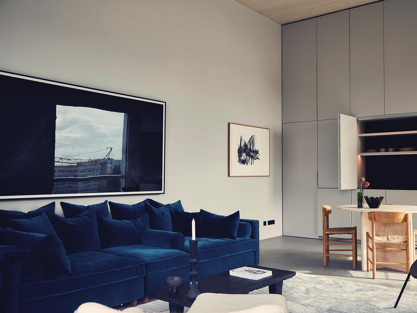 Helles Wohnzimmer mit samtigem blauen Sofa, offenem Küchenregal und Holztisch mit Stühlen.