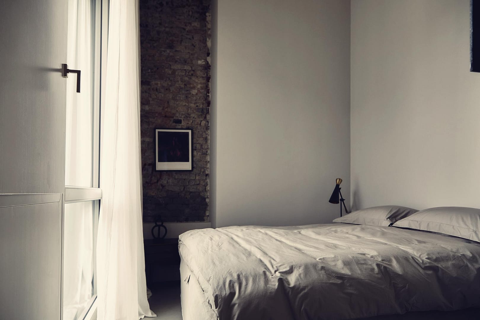 stadtflucht-homes-berlin-penthouse-522-schlafzimmer-bett-beistelltisch Schlafzimmer in einem Loft mit einem großen Bett mit grauem Stoffbezug, hellgrauer Bettwäsche und einem Beistelltisch mit einer schwarzen Lampe. Im Hintergrund ist eine Wand mit sichtbarem Backstein zu sehen. Ein Vorhang hängt neben einem Fenster.