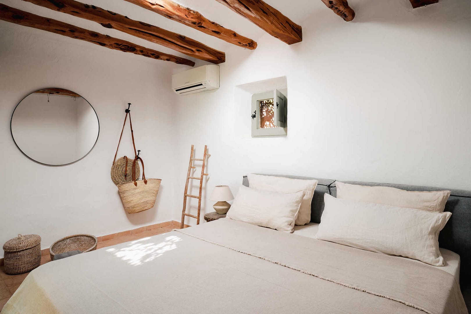 Schlafzimmer mit weißer Wand, Bett mit beigem Kopfteil und mehreren weißen Kissen, kleiner Holztisch mit Blumen und rundem Spiegel an der Wand