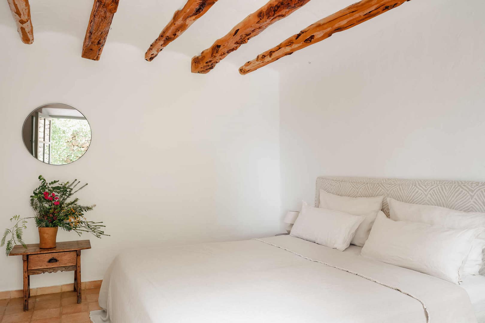 Schlafzimmer mit weißer Wand, Bett mit beigem Kopfteil und mehreren weißen Kissen, kleiner Holztisch mit Blumen und rundem Spiegel an der Wand