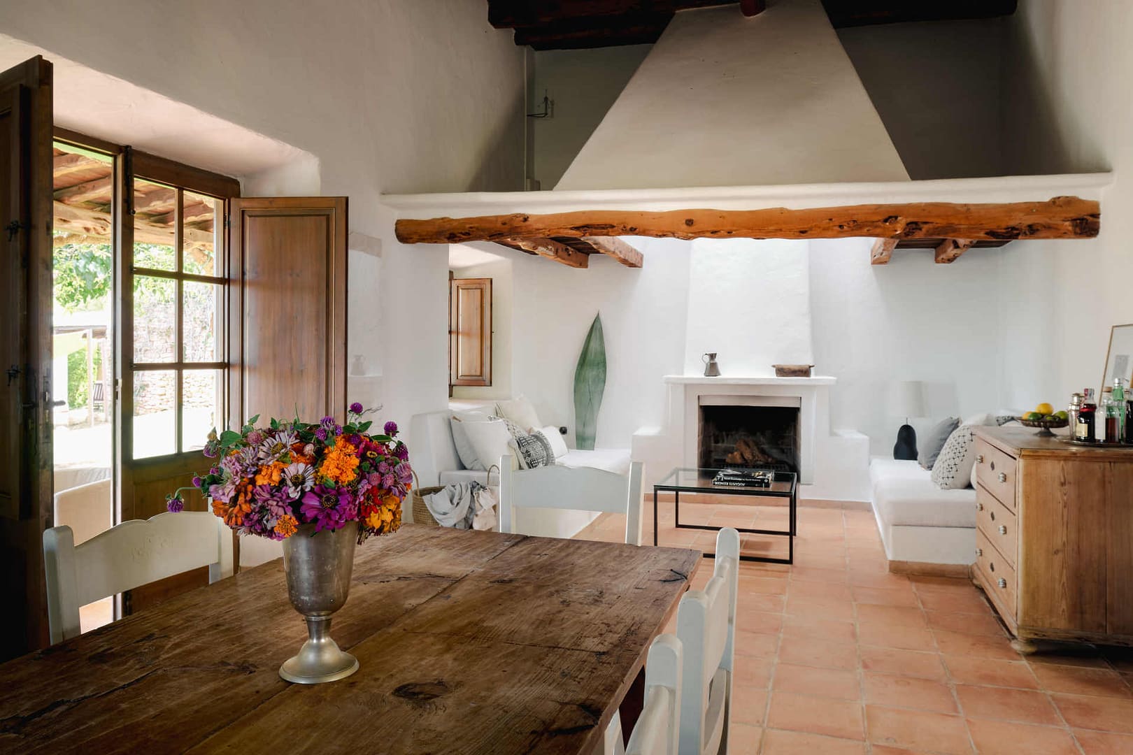 Heller Wohnraum in einer Ibiza-Finca mit Holzbalkendecke, Kamin, rustikalem Holztisch und weißen Möbeln