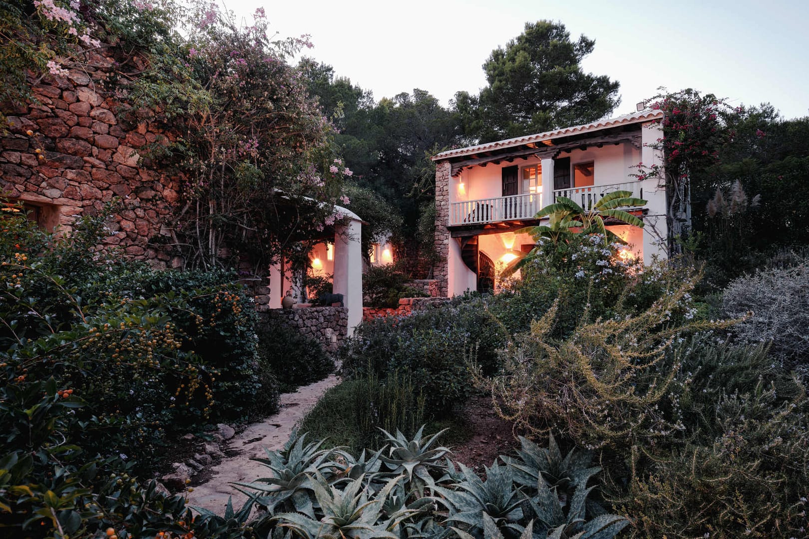 Ibiza-Finca mit Blick auf das Gästehaus, ein Garten bei Abendlicht mit mediterraner Pflanzenwelt und beleuchteten weißen Gebäuden