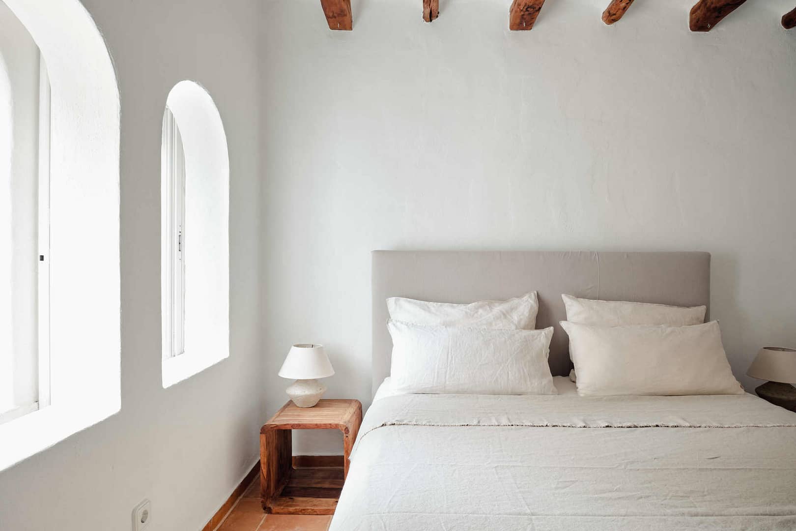 Schlafzimmer in einer Ibiza-Finca mit minimalistischem Design, hellen Wänden, einem großen Bett mit weißen Leinenbettwäsche und Holztischchen