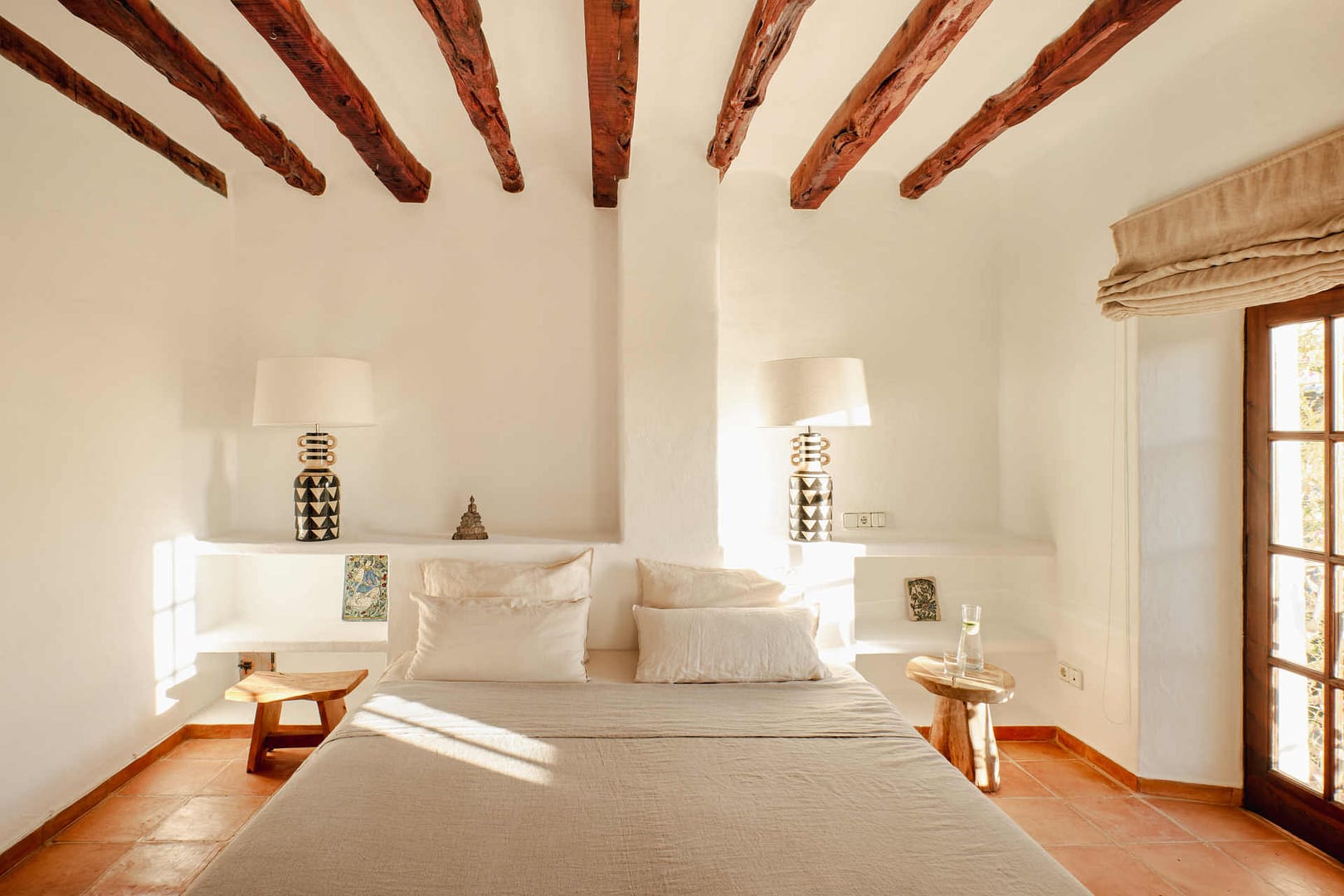 Schlafzimmer mit beigem Bett, Holzbalkendecke, zwei Nachttischen aus Holz, weißen Wänden und Fenstertür mit Holzrahmen