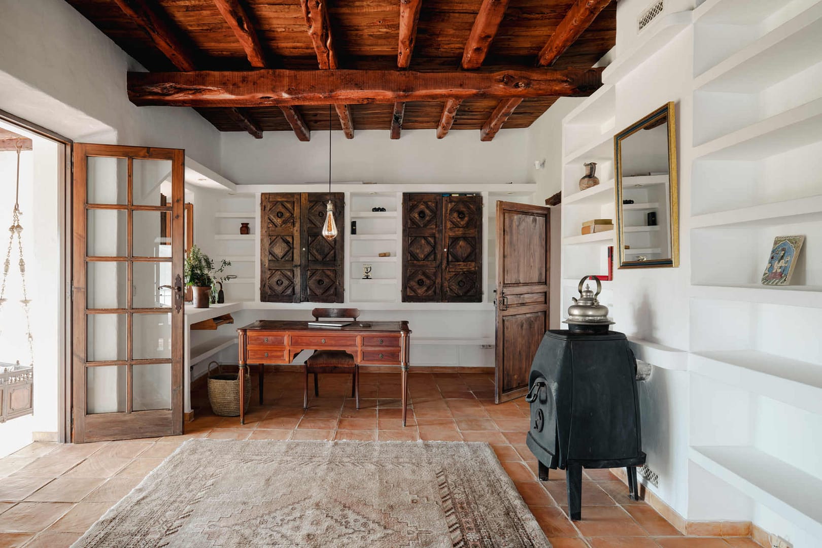 Wohnzimmer mit Holzbalkendecke, Holztür, offener Glasflügeltür, rotem Holztisch, Holzofen und weißen Regalen mit antiken Holztüren