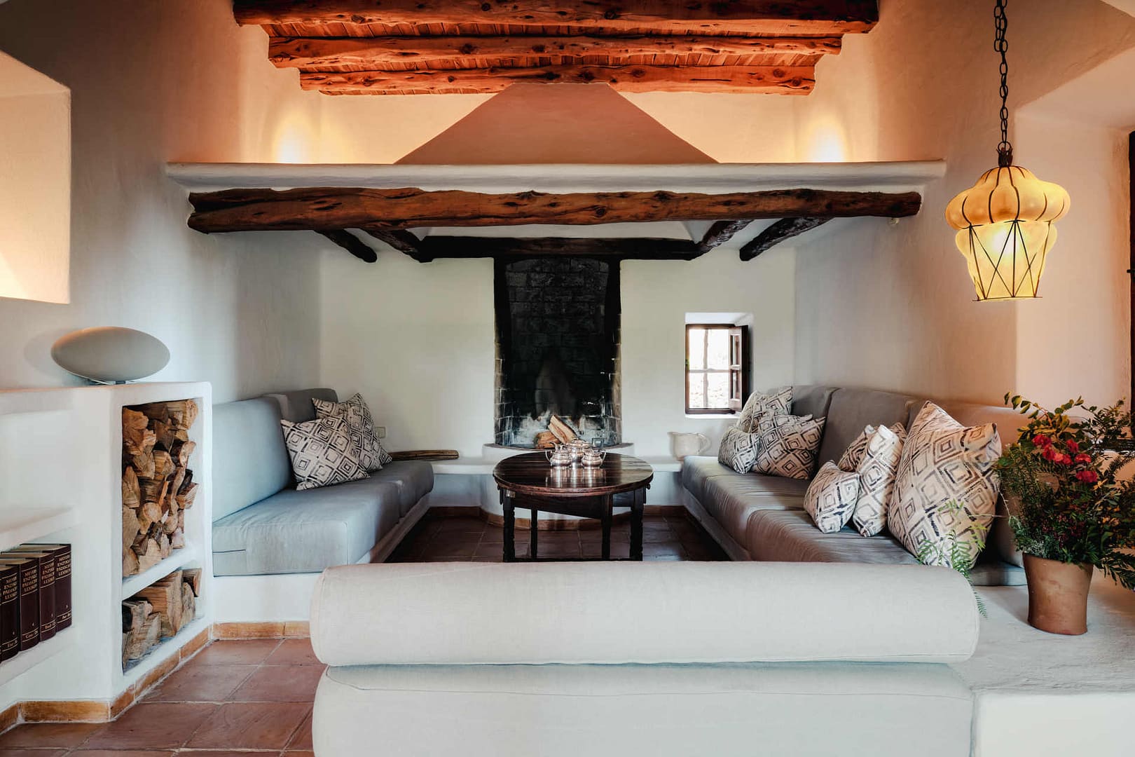 Wohnzimmer mit Kamin, hellgrauen Sofas mit gemusterten Kissen, runden Holztisch, Holzbalkendecke und Holzregal mit Kaminholz