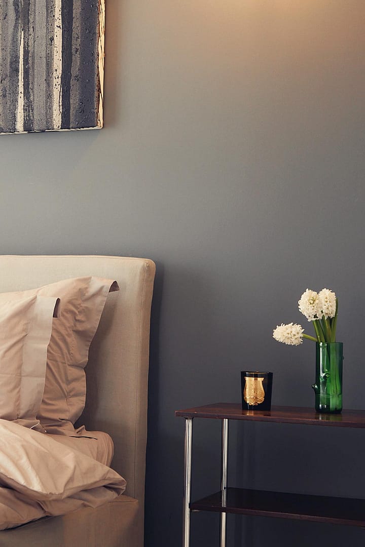 Nachttisch mit grünem Glasvasen mit weißen Blüten und Duftkerze neben einem hellen Bett vor grauer Wand im stilvollen Design-Studio Berlin