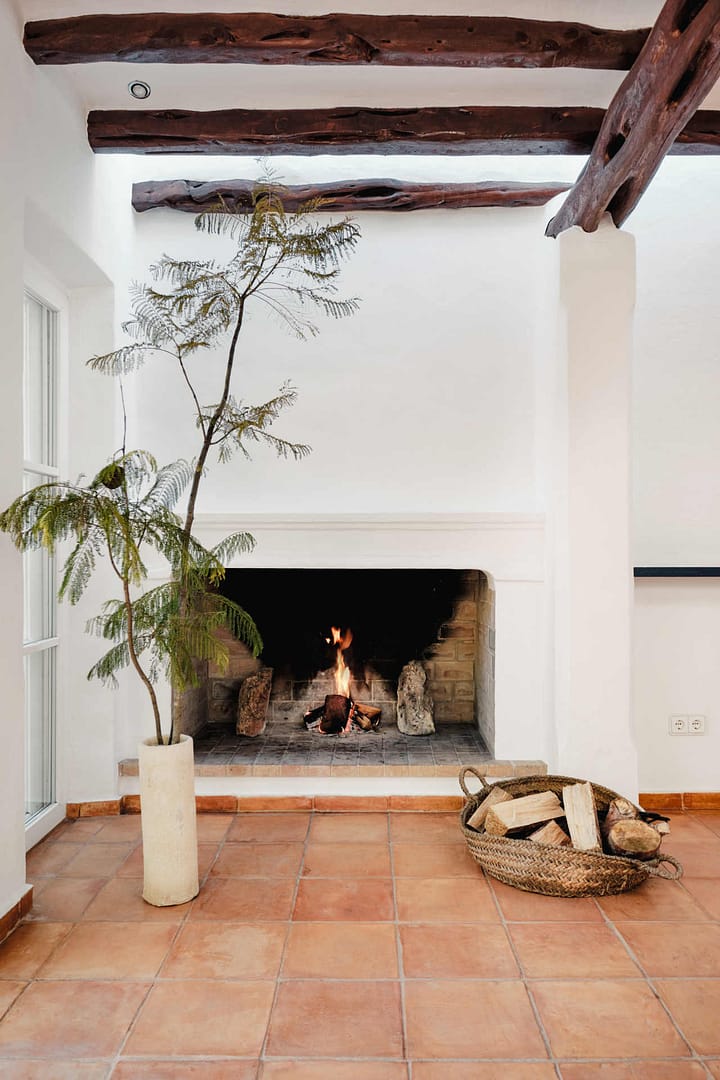 Brennender Kamin mit Holzkorb und Vase mit Baumzweig im Raum mit Terrakottafliesen.