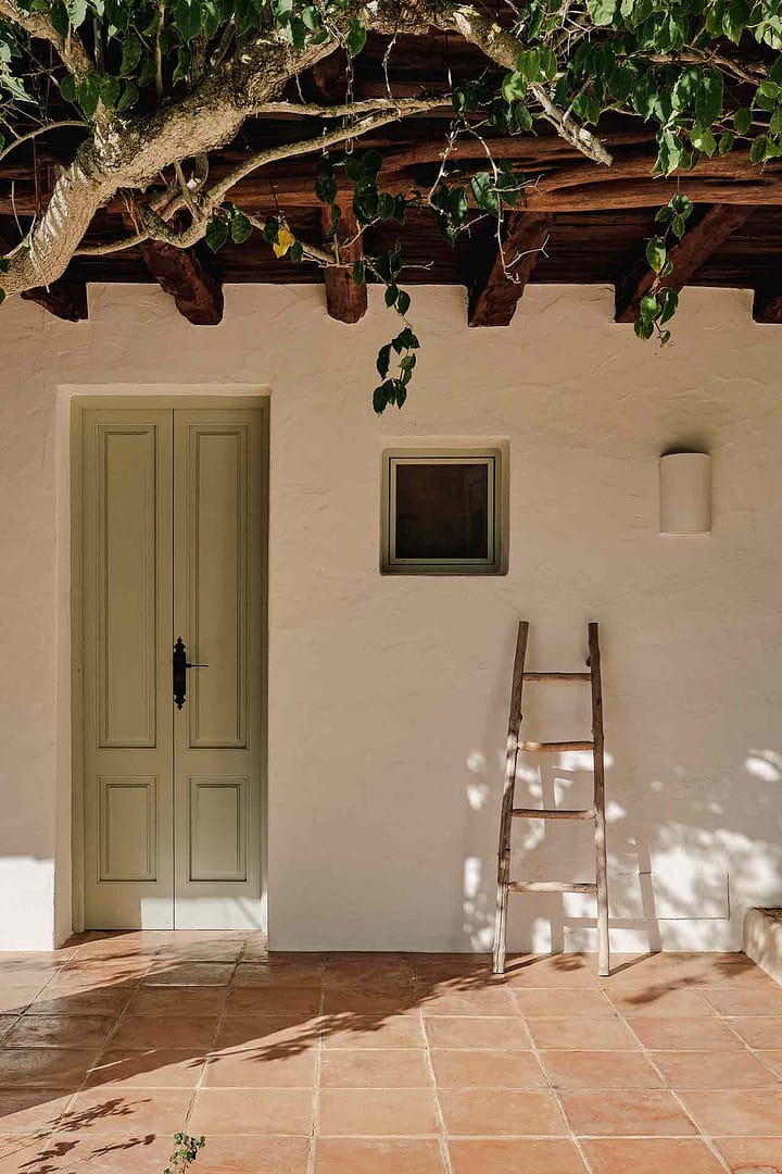 Eingang mit grüner Tür und kleinem Fenster an weißer Wand, daneben eine hölzerne Leiter, Terrakotta-Boden und Schatten von Ästen.