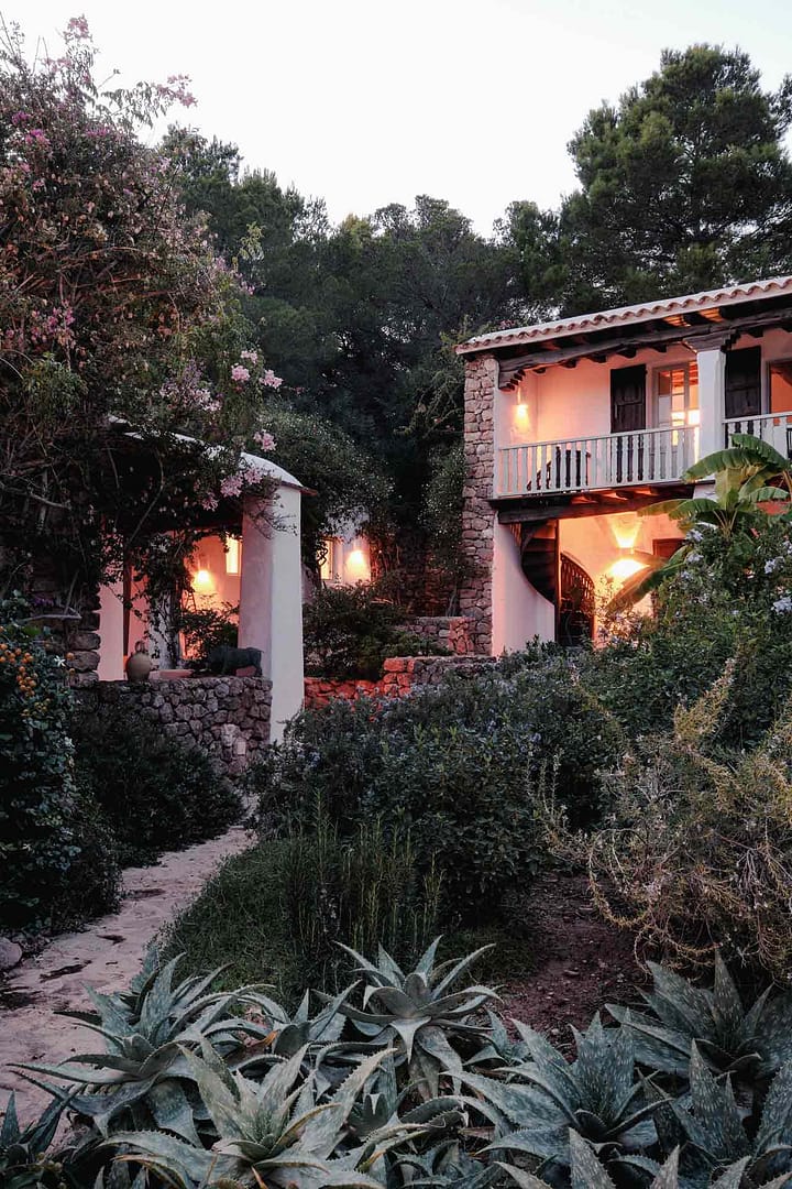 Terrasse einer Finca mit bepflanztem Garten, beleuchteten Mauern und Bäumen im Dämmerlicht.