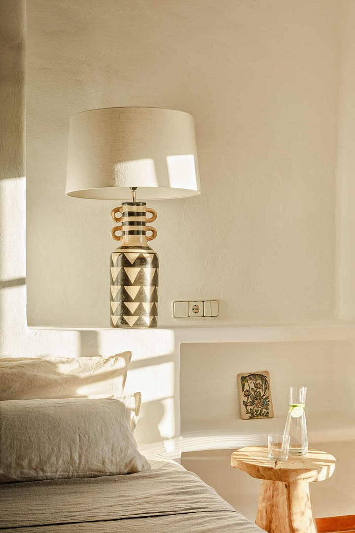 Schlafzimmer mit einem großen Bett, Nachttisch, Lampe mit gemustertem Fuß und einem kleinen Kunstobjekt in einer Nische.