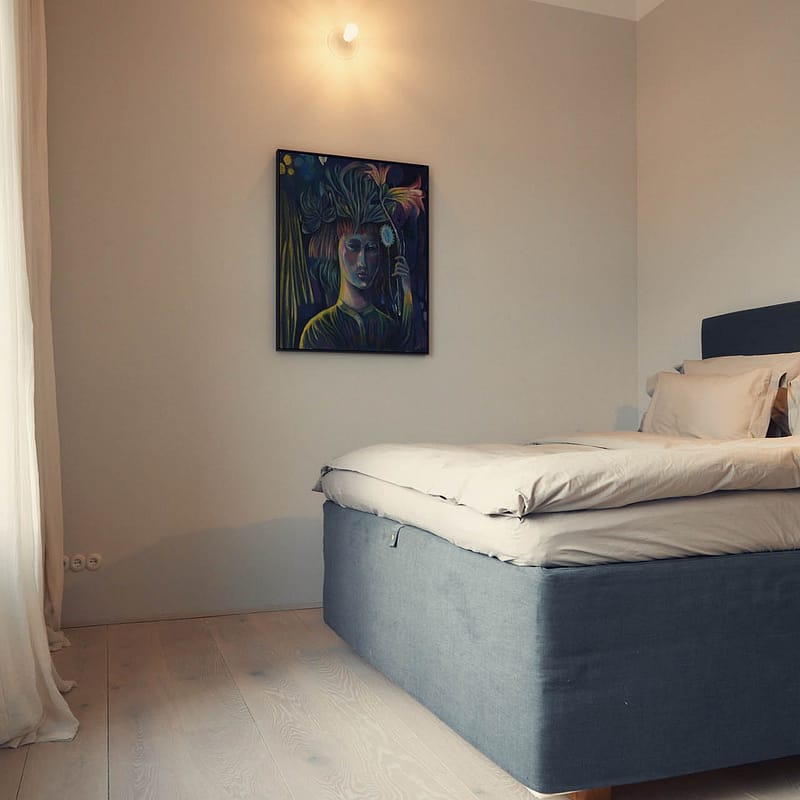 Schlafzimmer mit großem Bett, hellem Holzboden, hellem Vorhang und einem farbintensiven Bild an der Wand. Natürliches Licht fällt von links ins Zimmer.