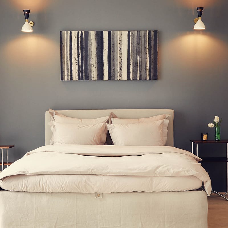 Modernes Schlafzimmer mit großem Bett vor einer grauen Wand, an der zwei Bilder hängen. Ein Nachttisch mit grüner Vase und weißen Blumen steht rechts neben dem Bett.