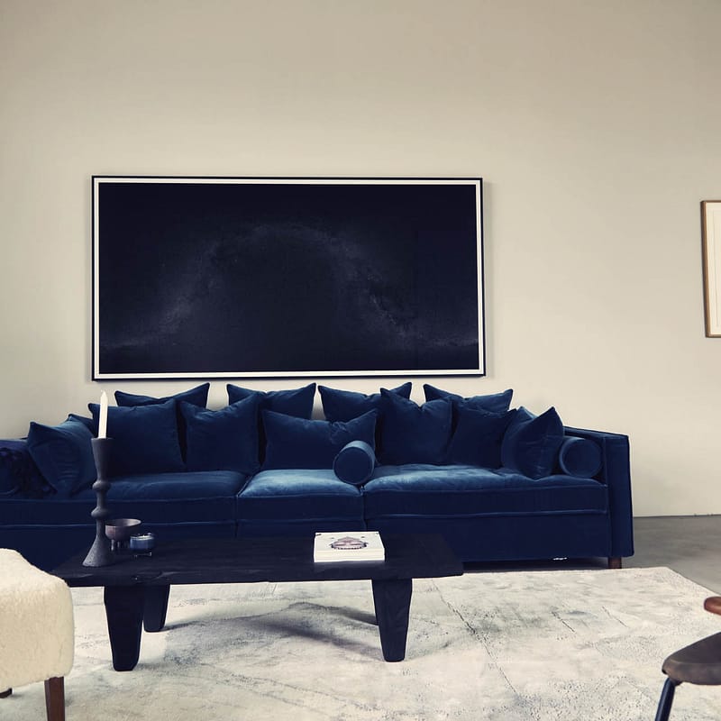 Wohnzimmer mit dunkelblauem Samtsofa, schwarzem Holztisch, einem weißen Fellhocker und mehreren Kissen. Im Hintergrund ein helles Fenster und zwei gerahmte Bilder an der Wand.