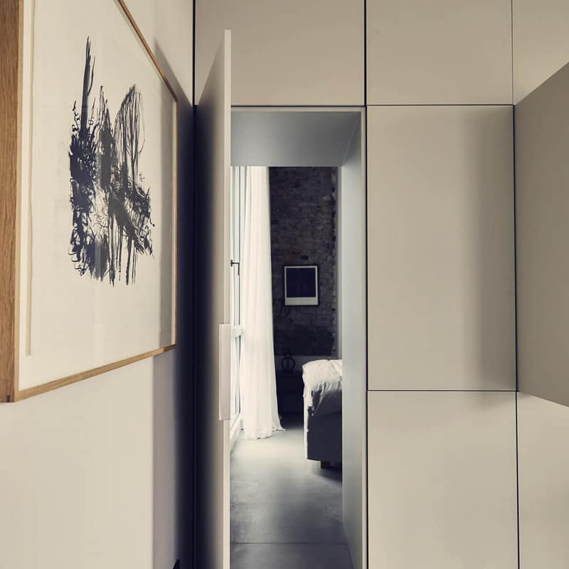 Blick durch schmalen Flur mit weißen Schränken rechts, einem Kunstbild links und durch die offene Tür ins Schlafzimmer mit Sichtziegelwand und weißen Vorhängen.