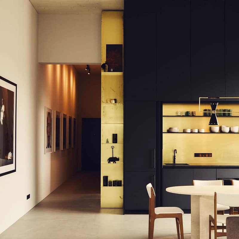 Moderner Essbereich mit ovalem Tisch und weißen Stühlen vor einer geöffneten Küchenzeile mit schwarzen Schränken und goldfarbenem Innenraum. Links ein heller Flur mit Bildern an der Wand.