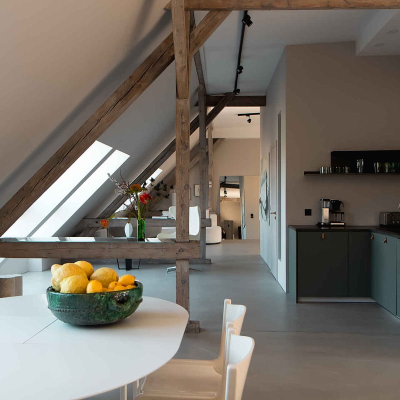 Design Studio Berlin – offene Küche mit lindgrünen Fronten, Obstschale auf Esstisch und Blick in den lichtdurchfluteten Wohnbereich mit Holzbalken