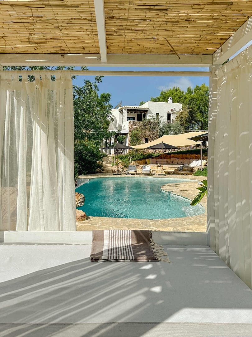 Blick vom Daybed unter einem Holz- und Bambusdach auf einen Pool, im Hintergrund weiße Finca und Garten.