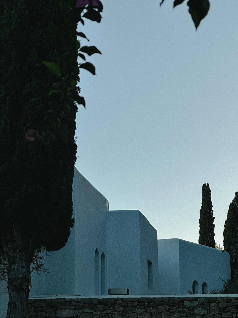 Weiße, kubische Finca-Fassade mit zwei schmalen Zypressen und Steinmauer im Vordergrund, fotografiert in der Abenddämmerung
