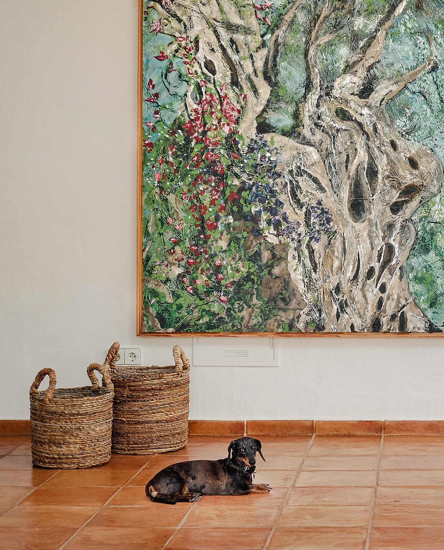 Kleiner Hund liegt auf terrakottafarbenem Fliesenboden vor zwei geflochtenen Körben und einer großen farbigen Wandkunst
