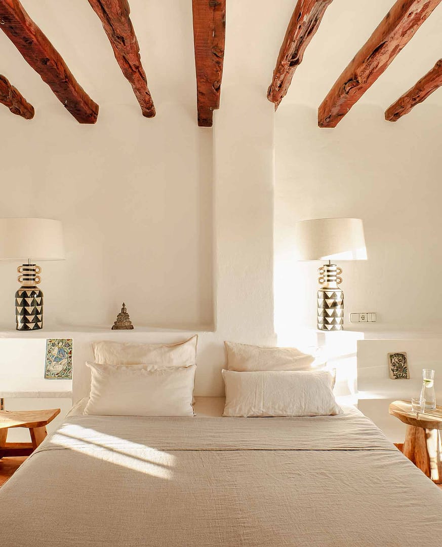 Schlafzimmer mit großem Bett, beigen Kissen, zwei Holztischen und zwei dekorativen Lampen sowie dekorativen Holzbalken an der Decke